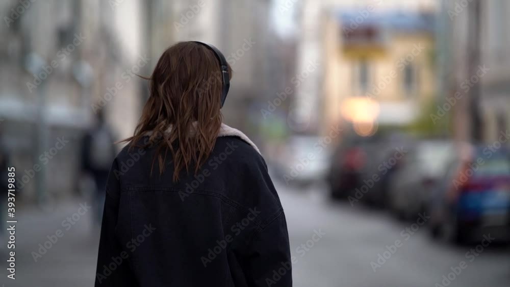 woman is walking in city at cold autumn day, listening to music by headphones, rear view in movement woman is walking in city at cold autumn day, listening to music by headphones, rear view in movement