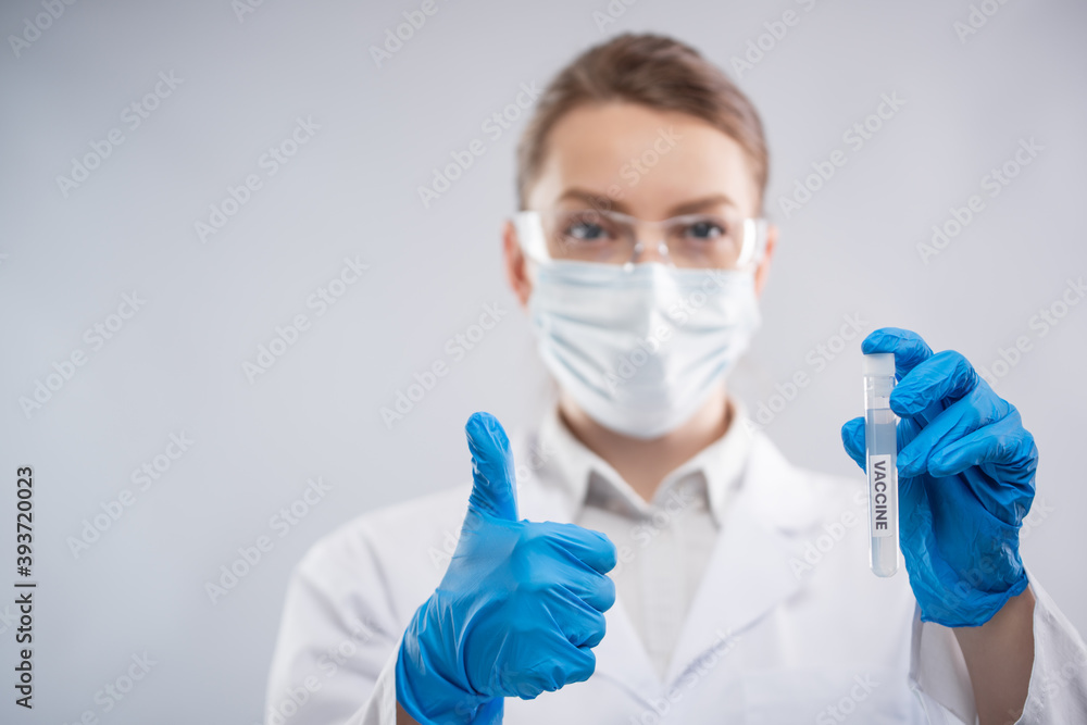 Close-up view of female microbiologist in biohazard protective clothing holds biological tube with vaccine and shows ok. Research and and development concept. Close-up view of female microbiologist in biohazard protective clothing holds biological tube with vaccine and shows ok. Research and and development concept.