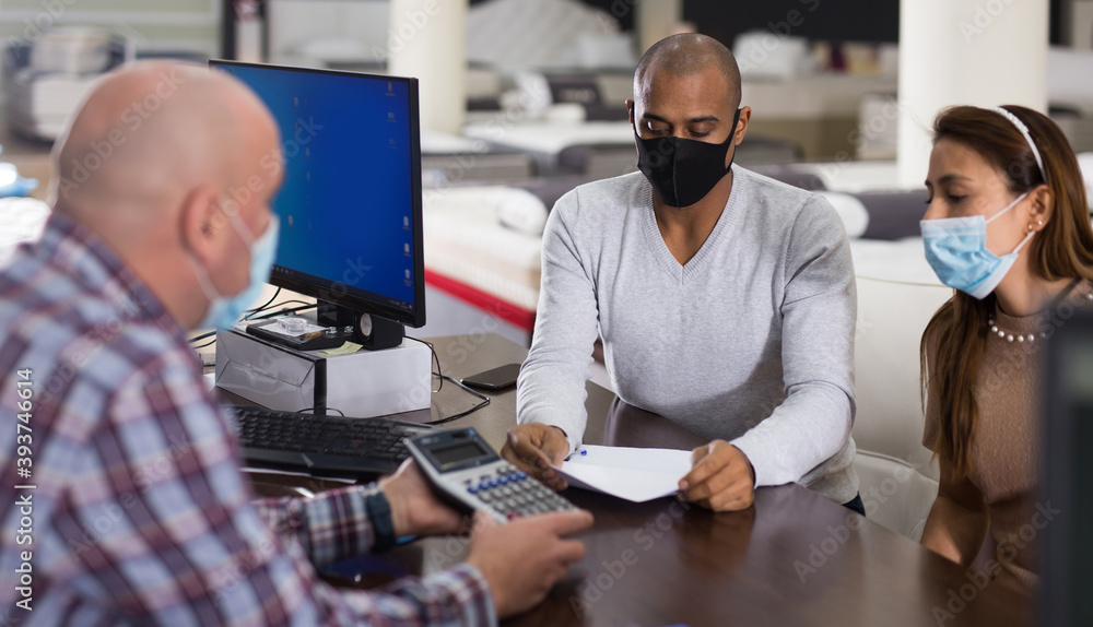 Fototapeta premium Young Hispanic couple in protective masks consulting with salesman when choosing new mattress in furniture store. Concept of shopping precautions and social distancing in pandemic