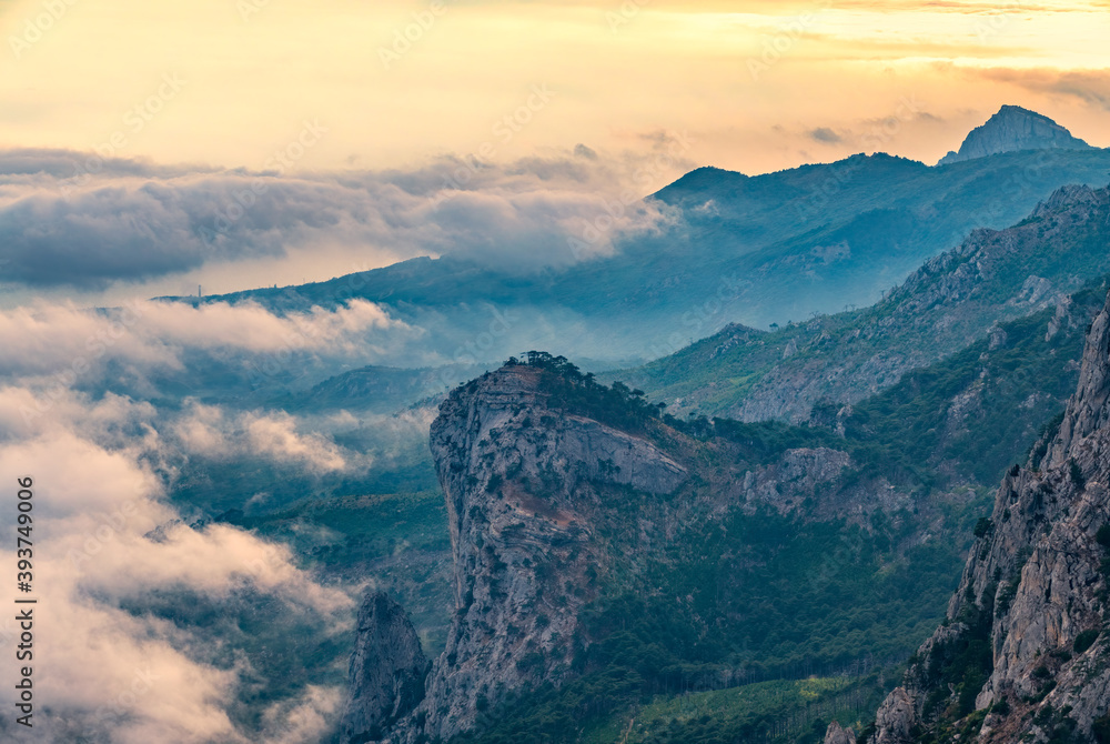 Obraz premium A majestic view of the rocky mountains and the valley in fog and clouds. Creamy fog covered the mountain valley in sunset light. Picturesque and gorgeous scene. Misty sunset over Crimea Mountains