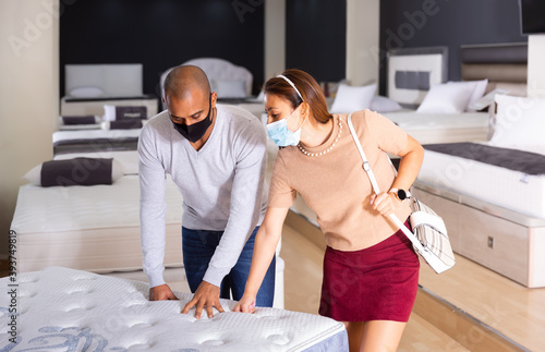 Young hispanic couple wearing protective face masks looking for new mattress for bed in furniture showroom. New lifestyle during coronavirus pandemic