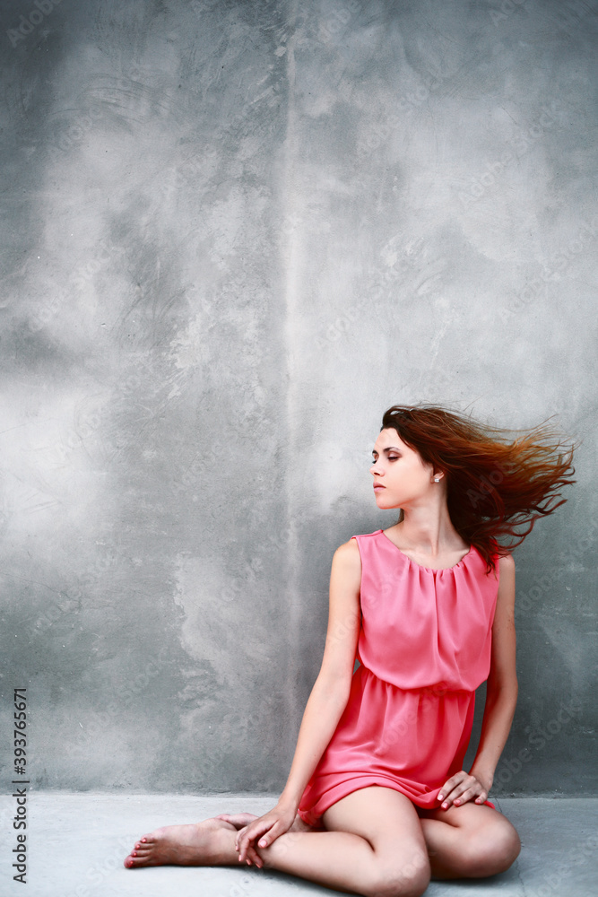 Obraz premium Young caucasian woman in a pink dress sitting near a concrete wall barefoot at sunset