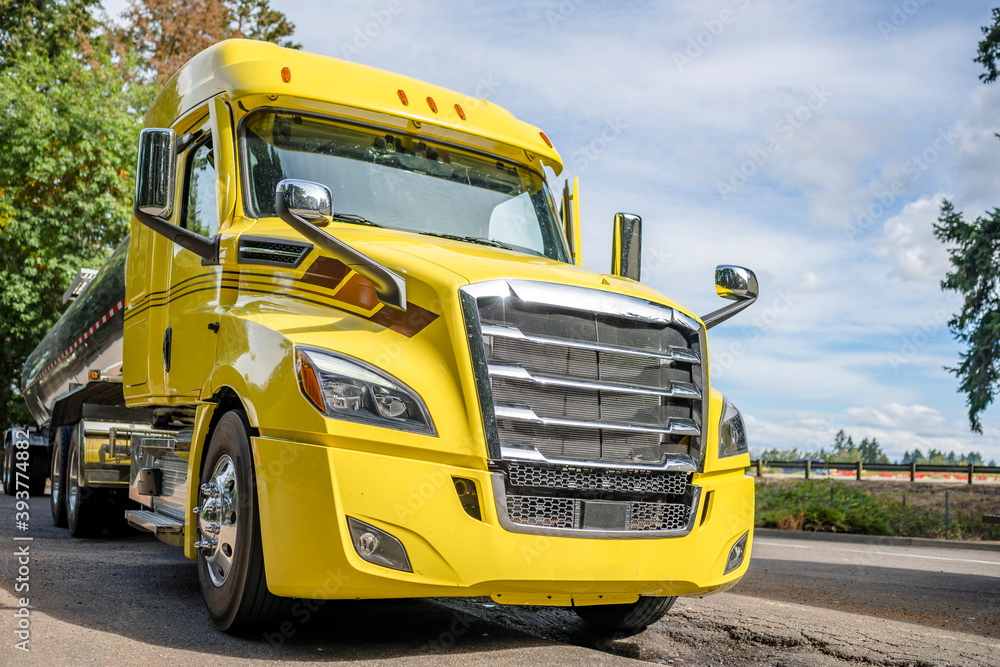Big rig yellow semi truck with shiny tank semi trailer standing on rest area parking lot with old trees Big rig yellow semi truck with shiny tank semi trailer standing on rest area parking lot with old trees