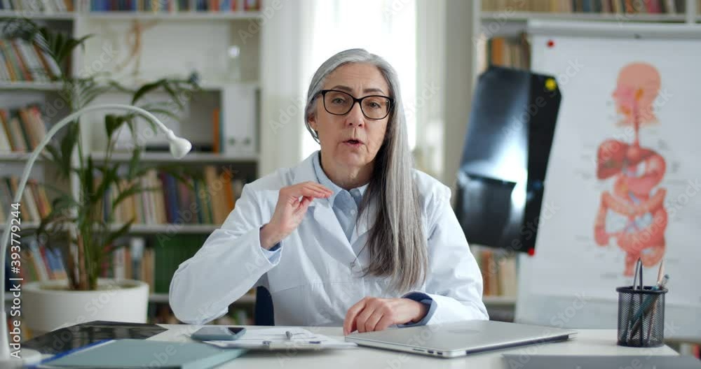 Cheerful elderly woman in glasses and white professional coat talking and gesturing while having online consultation. Female smiling doctor looking to camera in medical office Cheerful elderly woman in glasses and white professional coat talking and gesturing while having online consultation. Female smiling doctor looking to camera in medical office