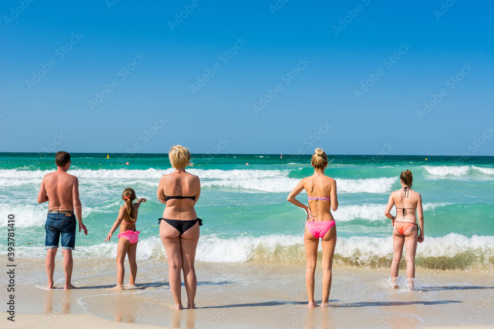 A group of tourists wearing swimsuit and bikini , and watching the waves, water splash flushing beach at Jumeirah beach at Persian gulf in Dubai, United Arab Emirates.
