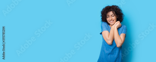 Adorable woman is posing on a blue studio wall gesturing pleasure and smiling near free space