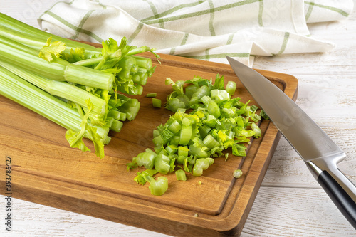 Cutting bunch of fresh green celery stalks into small pieces with a sharp knife on a brown wood cutting board over kitchen table. Natural antioxidant, healthy eating and weight loss diets ingredient.
