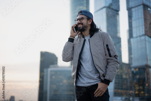 Attractive brunette man in a cap with glasses in a big city talking on a mobile phone