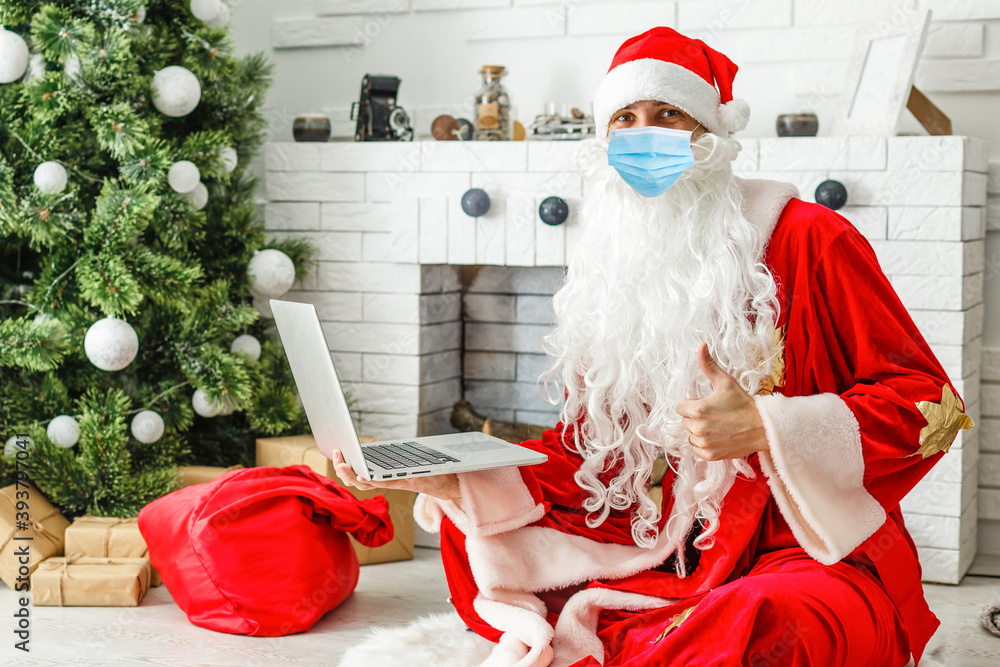 Obraz premium Santa Claus wearing face mask, holding gift box on xmas eve sitting at cozy home using laptop computer. Merry Christmas Covid 19 coronavirus social distance concept.