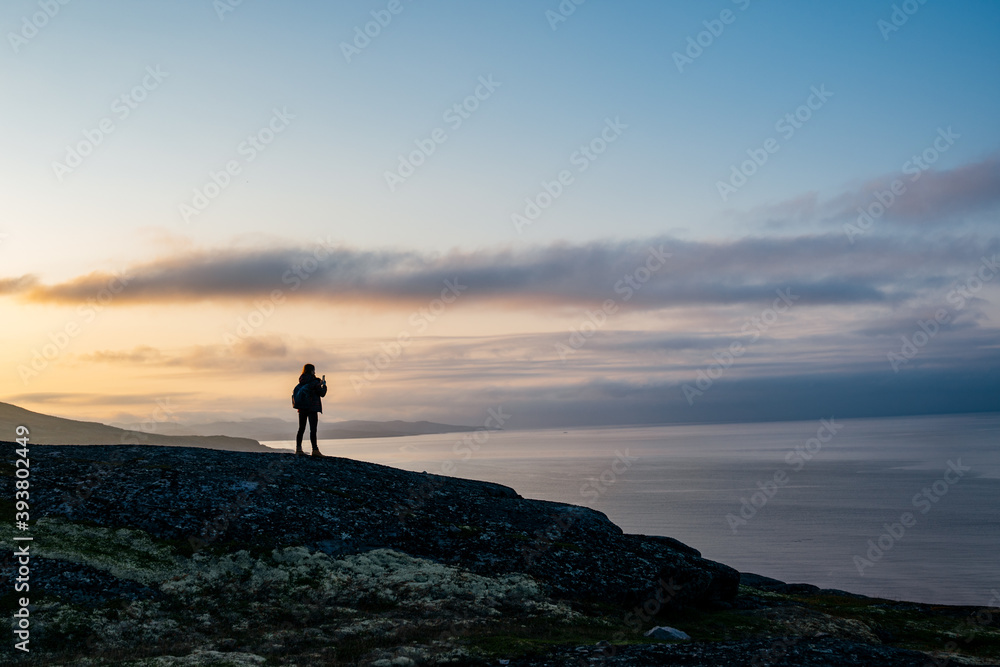Silhouette figure of young woman traveller enjoying the Beaty of nordic nature on the mountain near the sea taking the picture of the sunset 