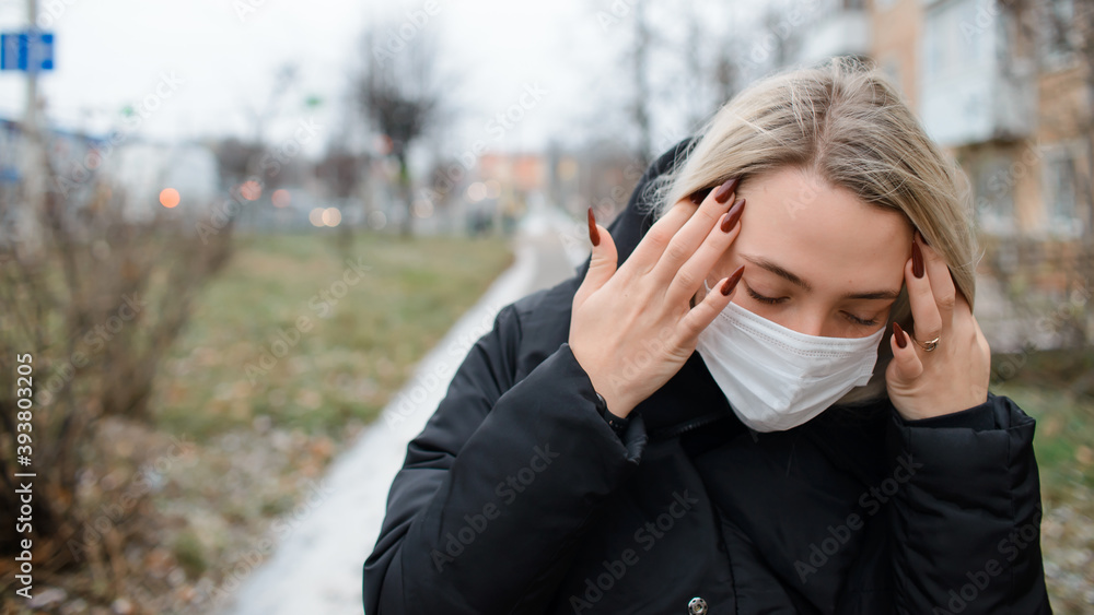 Young woman in a medical face mask outside. Girl holding her head. Concept of headache, depression, adolescent loneliness epidemic Young woman in a medical face mask outside. Girl holding her head. Concept of headache, depression, adolescent loneliness epidemic