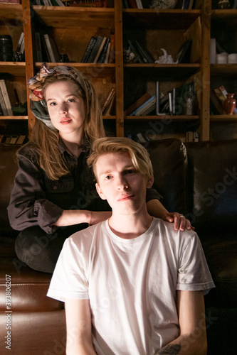 a young couple are sitting on a couch in a dark room, in the background there is a large bookcase with books