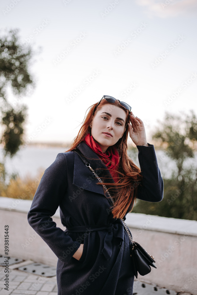 Pretty woman with green eyes and wide eyebrows and red hair posing on the city street  and smiling. Model dressed in warm clothes. Red scarf black coat and sunglasses. Romantic pretty female model