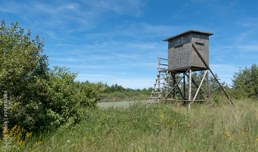 Geschlossener Hochsitz aus Holz auf einer wilden Wiese mit Büschen im Sommer Geschlossener Hochsitz aus Holz auf einer wilden Wiese mit Büschen im Sommer