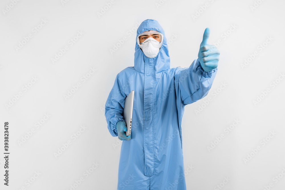 man doctor in a protective medical suit to protect against coronavirus, in a mask, glasses and gloves, with a laptop shows a thumb up, on a white background, healthcare man doctor in a protective medical suit to protect against coronavirus, in a mask, glasses and gloves, with a laptop shows a thumb up, on a white background, healthcare
