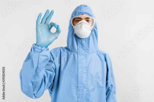 Positive man doctor, in protective blue suit, glasses, mask and gloves, shows okay gesture, on white background, healthcare concept, coronavrus