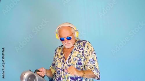 Crazy funny old caucasian man listening music using vintage stereo isolated on blue background dancing and having fun clubbing on vacation