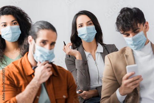 young hispanic businesspeople in medical masks listening lecture while man chatting on smartphone on blurred foreground