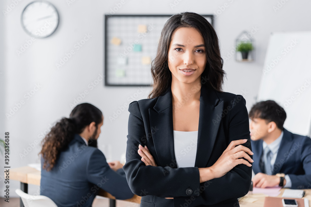 Confident businesswoman with crossed arms looking at camera with blurred co-workers on background Confident businesswoman with crossed arms looking at camera with blurred co-workers on background