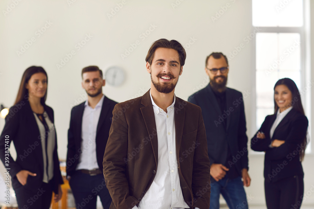 Smiling businessman or company owner looking at camera over positive colleagues at background Smiling businessman or company owner looking at camera over positive colleagues at background