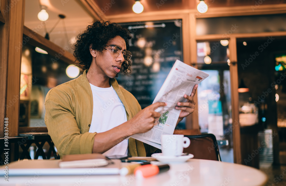 Serious handsome male 20s in trendy wear and eyeglasses enjoying leisure holding newspaper getting daily information, pensive businessman checking financial economy news from columns in print media