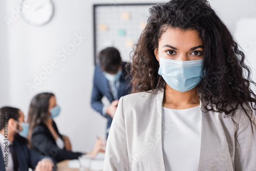 Portrait of curly hispanic businesswoman in medical mask looking at camera with blurred colleagues working on background