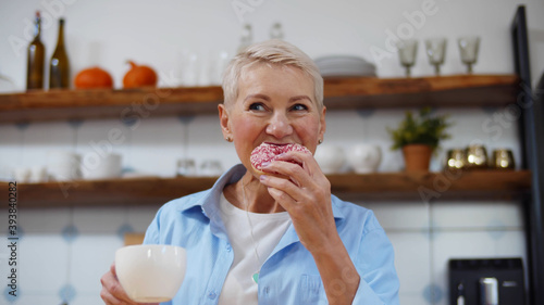 Smiling senior woman drinking coffee and eating delicious glazed donut at home