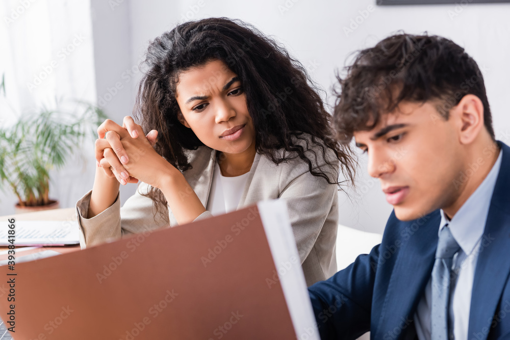 Obraz premium Pensive hispanic businesswoman looking at papers near colleague on blurred foreground in office