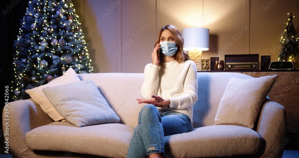 Obraz premium Happy Caucasian beautiful female in medical mask sitting on sofa at decorated home near xmas glowing tree in evening and calling on smartphone greeting friend with holidays. Christmas concept