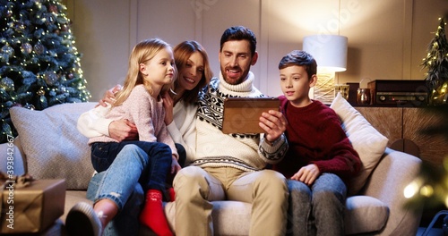 Caucasian cheerful family with children sitting at decorated home near glowing xmas tree and speaking on video chat online on tablet congratulate friends and relatives with holidays. Happy New Year