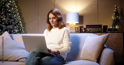 Photography Happy young beautiful Caucasian woman sitting on sofa at decorated home near glowing christmas tree in evening and typing and browsing on laptop on Xmas Eve