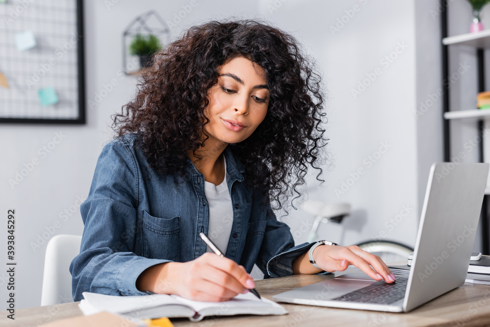 curly woman using laptop and writing in notebook while working from home curly woman using laptop and writing in notebook while working from home