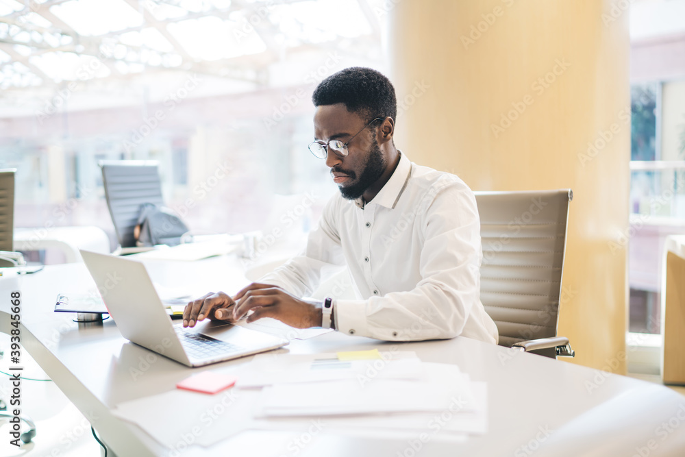 Dark skinned male entrepreneur in smart casual wear and spectacles typing on laptop computer share media and publication, serious man employee concentrated on job at working place in office