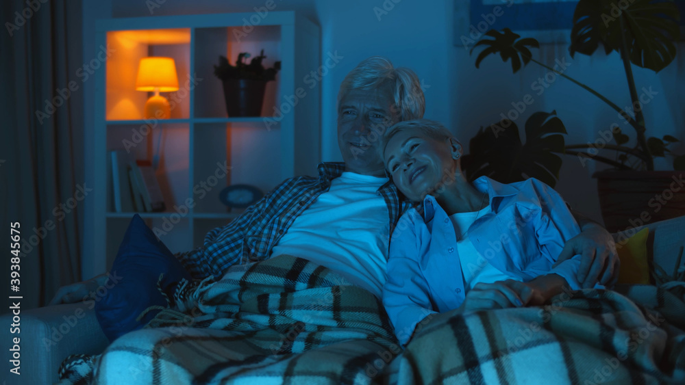 Fototapeta premium Modern senior couple hugging and watching tv together sitting on comfortable couch at home