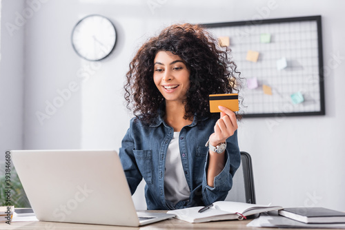 cheerful young woman holding credit card near laptop on desk