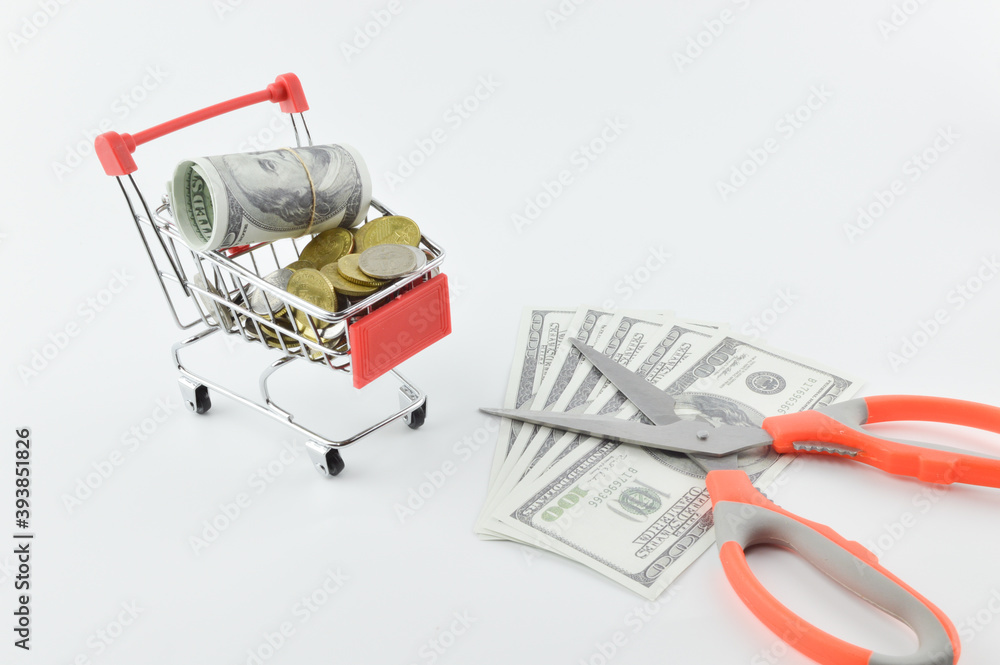 Selective focus of shopping cart, banknotes, coins and scissors on a white background. Devaluation concept.