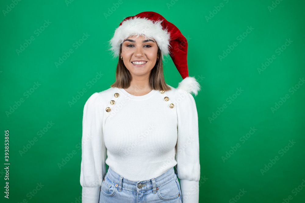 Young beautiful woman wearing a Santa hat and white sweater over green background with a happy face standing and smiling with a confident smile showing teeth Young beautiful woman wearing a Santa hat and white sweater over green background with a happy face standing and smiling with a confident smile showing teeth