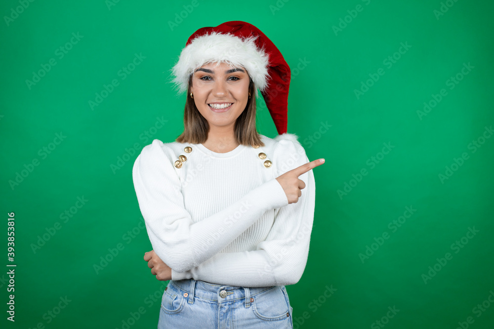 Young beautiful woman wearing a Santa hat and white sweater over green background smiling and pointing with hand and finger to the side Young beautiful woman wearing a Santa hat and white sweater over green background smiling and pointing with hand and finger to the side