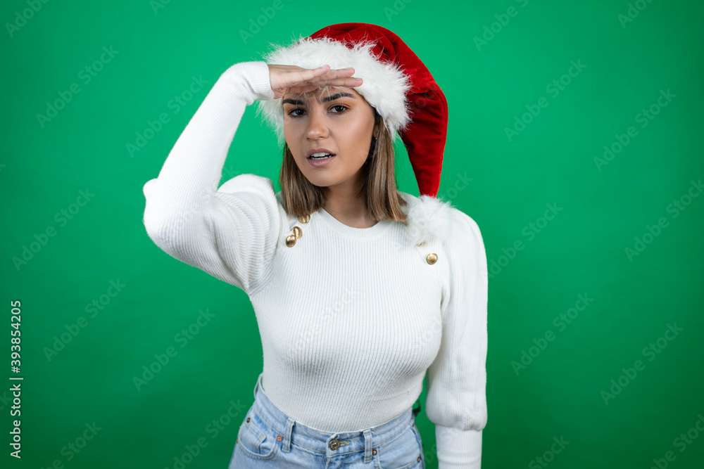 Young beautiful woman wearing a Santa hat and white sweater over green background very happy and smiling looking far away with hand over head. searching concept. Young beautiful woman wearing a Santa hat and white sweater over green background very happy and smiling looking far away with hand over head. searching concept.