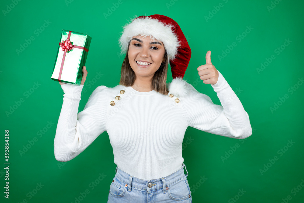 Young beautiful woman wearing a Santa hat holding a gift and white sweater over green background smiling and doing the ok signal with her thumb Young beautiful woman wearing a Santa hat holding a gift and white sweater over green background smiling and doing the ok signal with her thumb