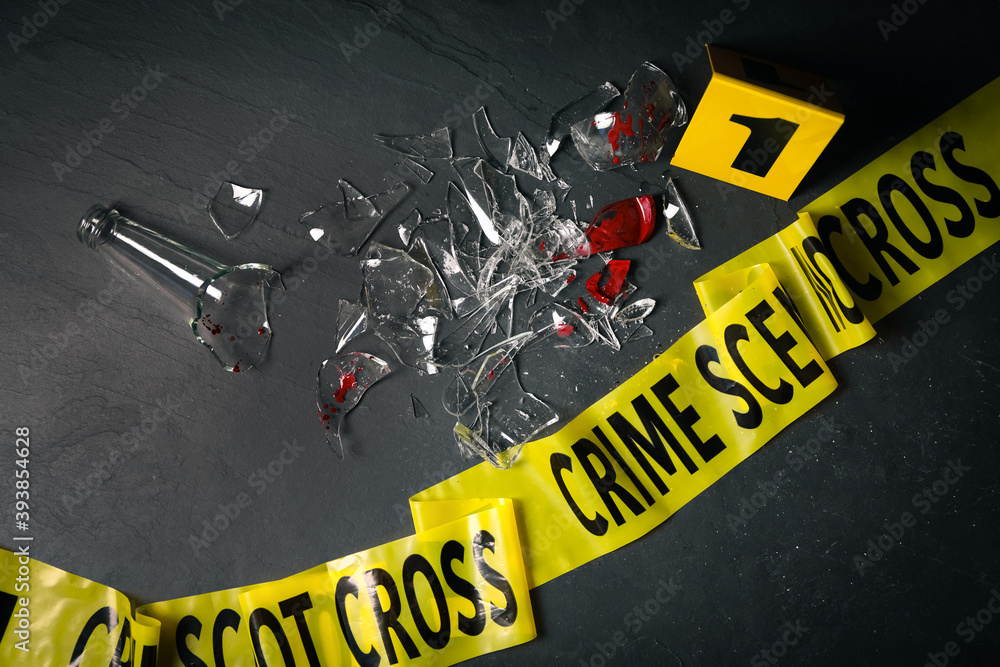 Fototapeta premium Broken bottle with blood, yellow tape and evidence marker on black slate background, flat lay. Crime scene