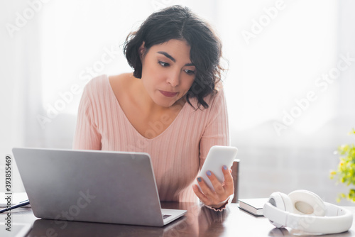 thoughtful hispanic woman looking at smartphone while sitting near laptop and wireless headphones