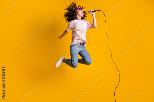 Photo portrait full body view of ecstatic woman jumping up singing into microphone isolated on vivid yellow colored background