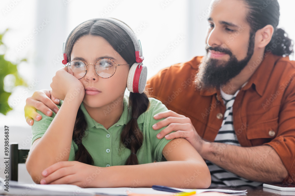 Positive hispanic father comforting sad daughter, while sitting at table with studying materials on blurred foreground Positive hispanic father comforting sad daughter, while sitting at table with studying materials on blurred foreground
