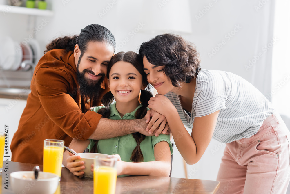 Hispanic parents hugging smiling daughter during breakfast on blurred foreground