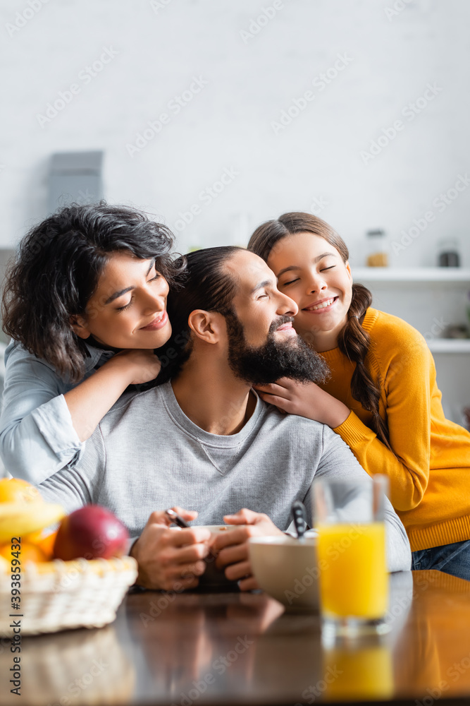 Smiling hispanic daughter and woman hugging father near breakfast on blurred foreground