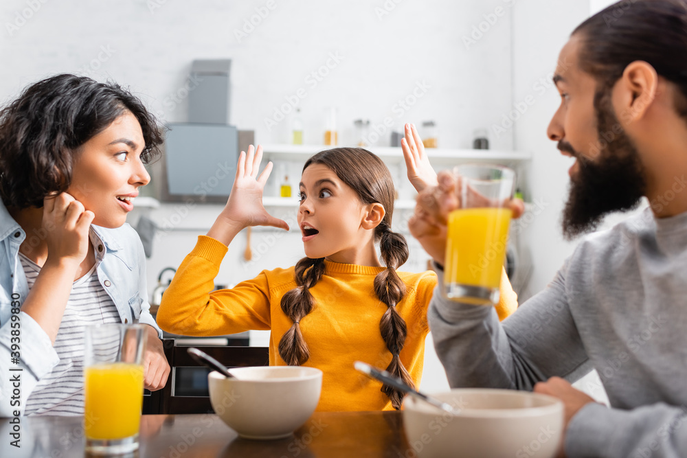 Excited girl talking to hispanic parents during breakfast on blurred foreground