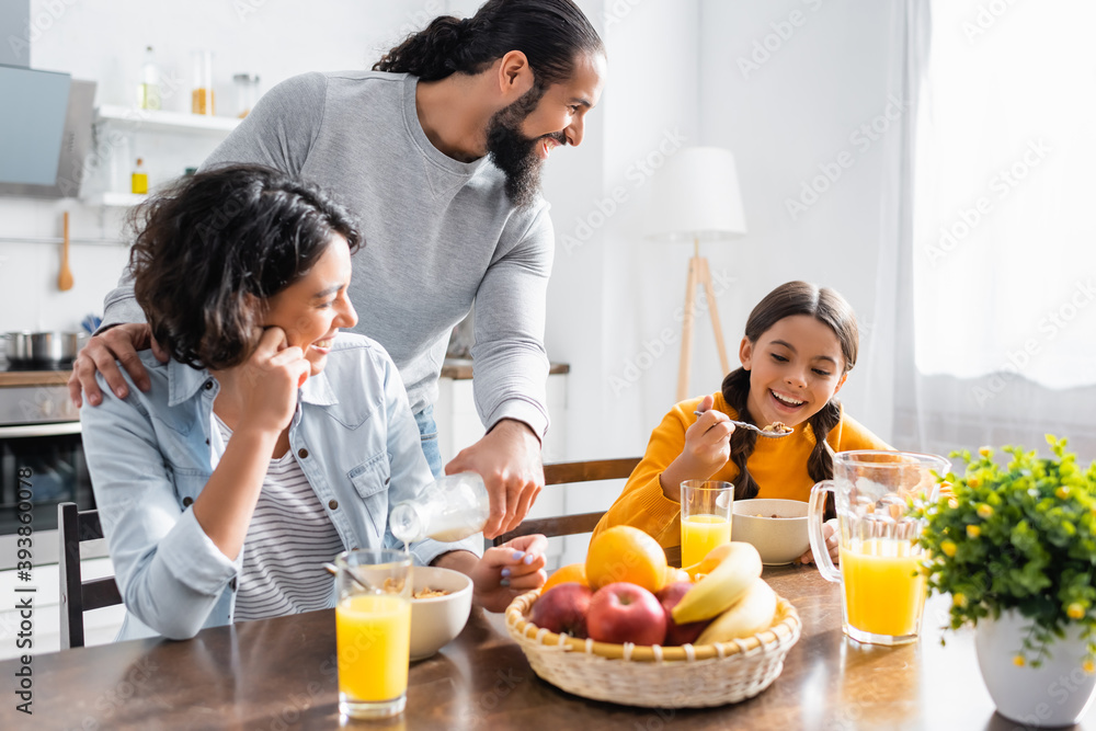 Hispanic man pouring milk near wife and smiling daughter with cereals in kitchen Hispanic man pouring milk near wife and smiling daughter with cereals in kitchen