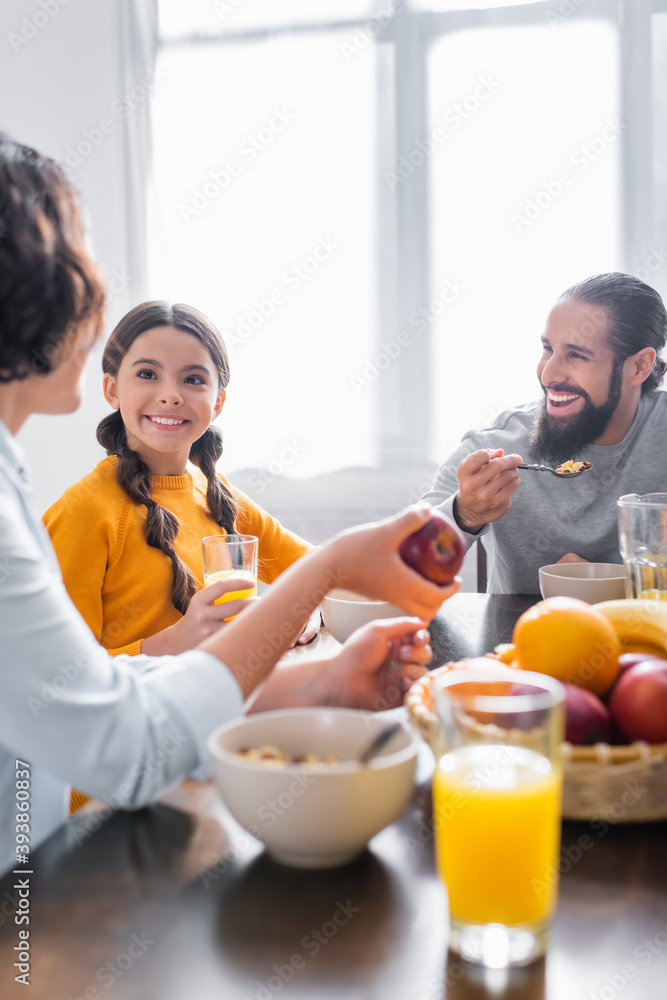 Smiling hispanic girl holding glass of orange juice near parents during breakfast Smiling hispanic girl holding glass of orange juice near parents during breakfast