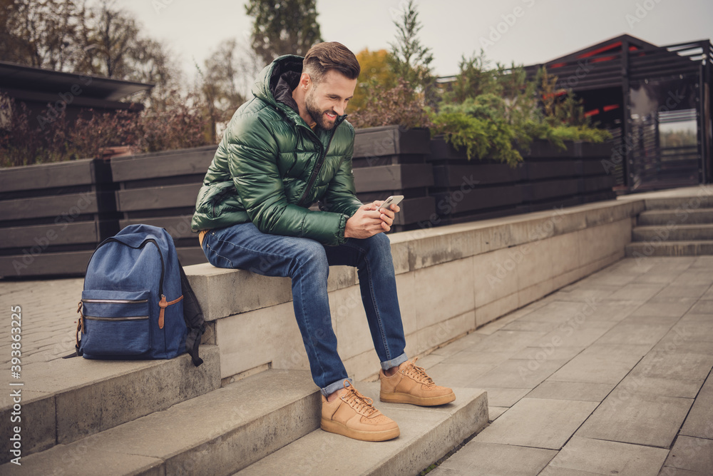 Profile side photo of attractive beaming young guy dressed green coat schoolbag having rest sitting stairs outdoors city street